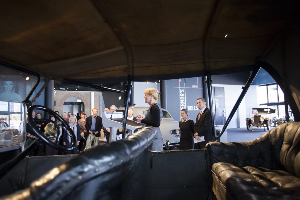 A woman is standing at a lectern addressing a small group of people in a museum exhibition area which has old cars lined up on display. Behind her is a man wearing a suit and a woman wearing a scarf. They standing are both near a Rolls-Royce limousine which has a crown instead of a number plate. The photographer has taken the image looking through an old car which has no windows but a canopy roof. The leather seats and steering wheel of the car can be seen. 